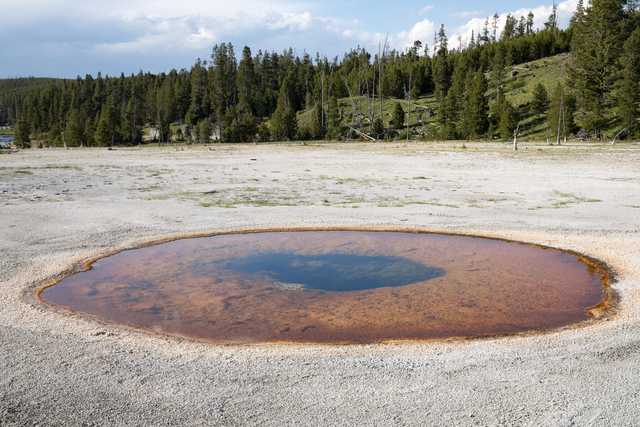 A circular, orange hot spring rests on a vast white and grey landscape, with a forest visible in the distance.