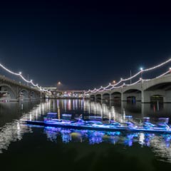 An illuminated boat glides across calm waters beneath a bridge adorned with festive lights at night.
