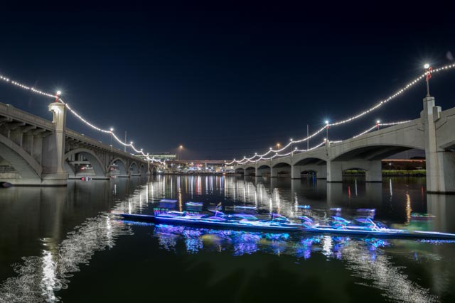 An illuminated boat glides across calm waters beneath a bridge adorned with festive lights at night.