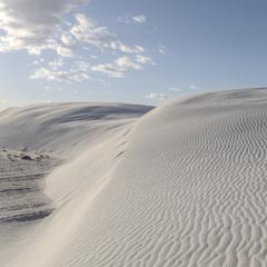 A series of undulating white dunes extends to a dark, rippled surface under a blue sky with scattered clouds.