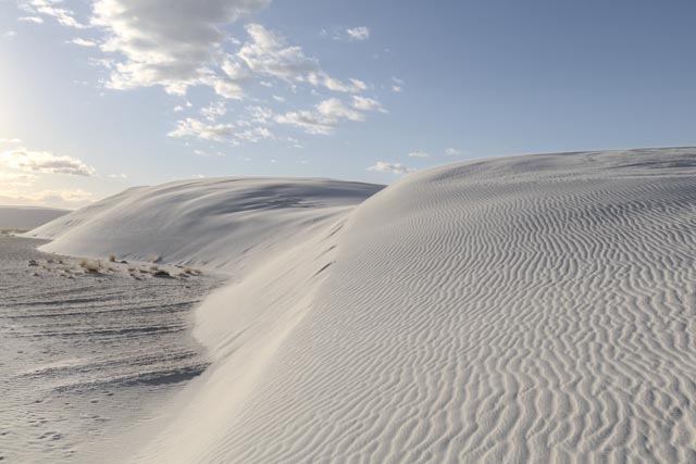 A series of undulating white dunes extends to a dark, rippled surface under a blue sky with scattered clouds.