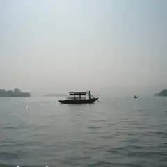 A silhouetted boat with a canopy floats on calm water, steered by a single figure, with tree-covered islands and hazy hills in the distance.