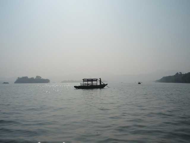 A silhouetted boat with a canopy floats on calm water, steered by a single figure, with tree-covered islands and hazy hills in the distance.