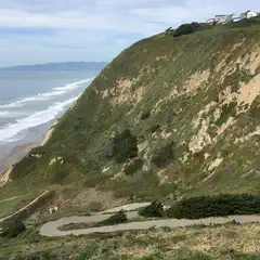 A winding paved road traverses a steep, grassy hillside overlooking a beach and ocean.