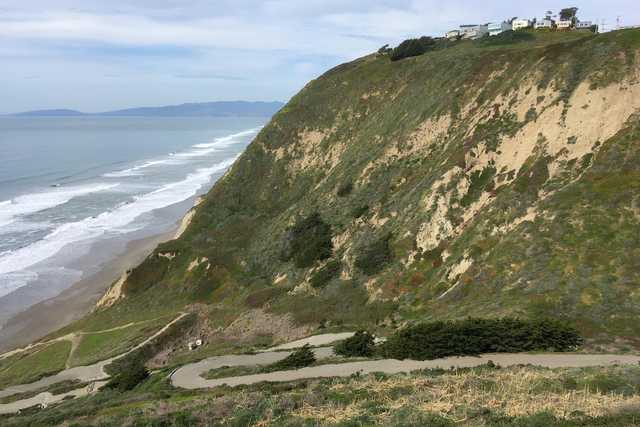 A winding paved road traverses a steep, grassy hillside overlooking a beach and ocean.