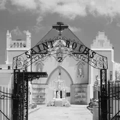 A black wrought iron gate with the words "Saint Roch's Campo Santo 2" arches over a paved pathway leading to a white chapel with a crucifix and statues.
