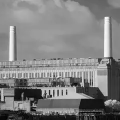 Four tall, fluted chimneys rise above a large, multi-story industrial building with a grid of windows, set against a cloudy sky.