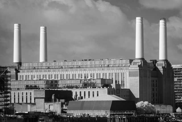 Four tall, fluted chimneys rise above a large, multi-story industrial building with a grid of windows, set against a cloudy sky.