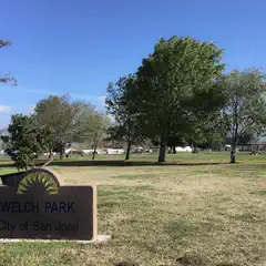 A sign labeled "Welch Park, City of San José" stands in a grassy field with trees and distant houses and hills.