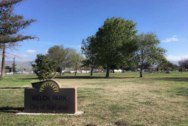A sign labeled "Welch Park, City of San José" stands in a grassy field with trees and distant houses and hills.