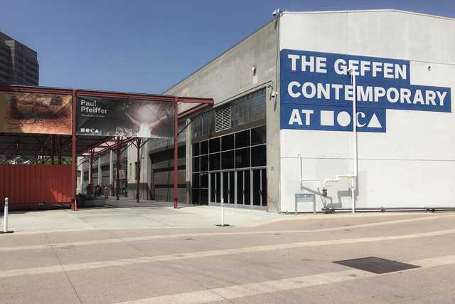 A modern industrial-style building displays a prominent blue sign with white text, alongside a red-framed structure with banners advertising an exhibition.
