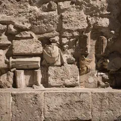 A weathered stone wall with embedded fragments of classical sculpture, including a draped bust and carved head, set between fluted columns.