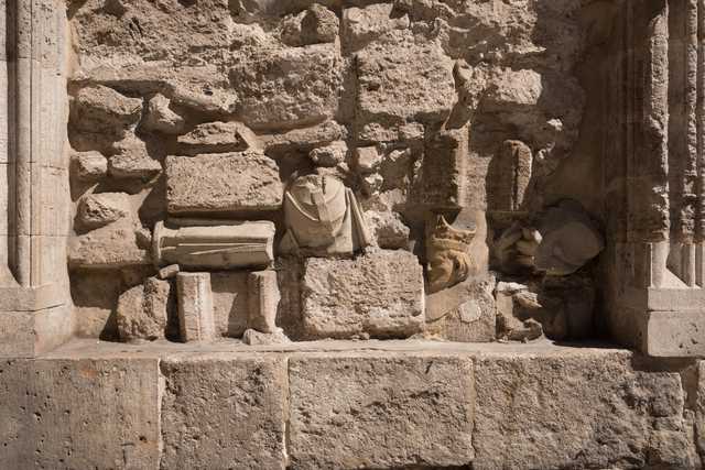 A weathered stone wall with embedded fragments of classical sculpture, including a draped bust and carved head, set between fluted columns.