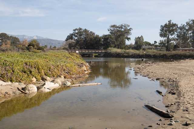 A shallow stream flows through a sandy channel, bordered by vegetation and rocks, with a small bridge crossing in the distance under a hazy sky.