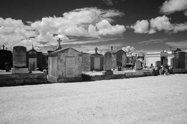 A row of mausoleums and gravestones occupies a grassy area beneath a cloudy sky.