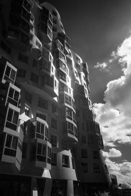A high-rise residential building features staggered, curved balconies and reflective surfaces under a partly cloudy sky.