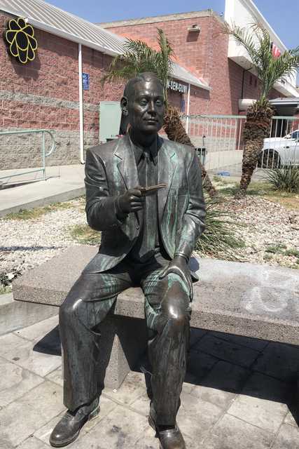 A statue of a man sitting on a bench, dressed in a suit and tie, with a book in his hand, located outside a building under a clear sky.