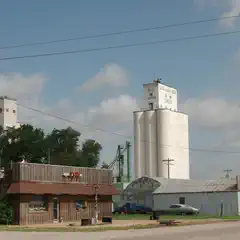 A wooden building with an awning stands next to two concrete grain elevators under a blue sky.