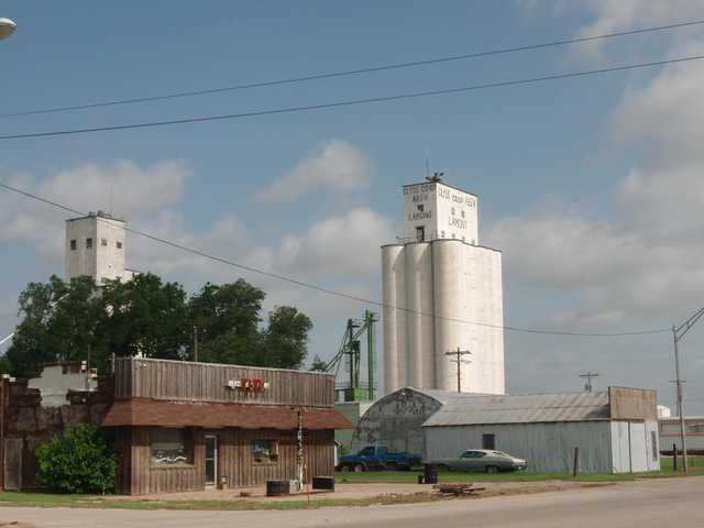 A wooden building with an awning stands next to two concrete grain elevators under a blue sky.