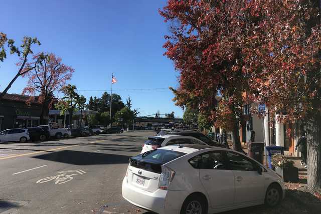 A street scene shows parked cars lining both sides of a road, with trees displaying autumn foliage and an American flag flying in the background.