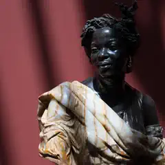 A carved figure of a woman with styled hair, a decorative headpiece, and a patterned garment, set against a red background.