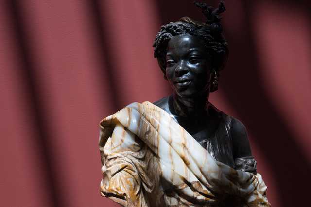 A carved figure of a woman with styled hair, a decorative headpiece, and a patterned garment, set against a red background.