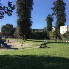 A sunny day at a park with a playground, trees, and people sitting on benches.