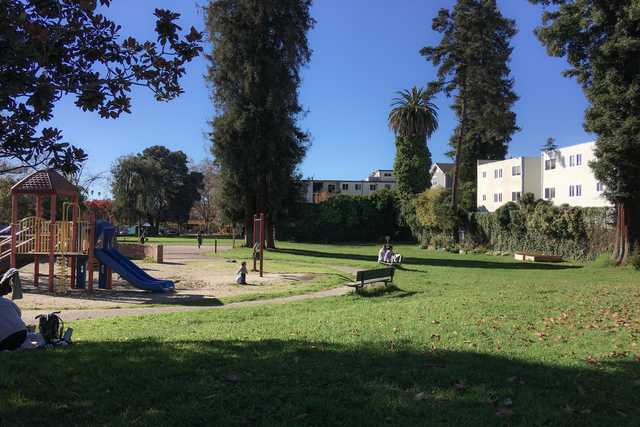 A sunny day at a park with a playground, trees, and people sitting on benches.