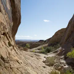 A narrow canyon with sandstone walls contains a dry streambed and leads to a distant, flat landscape under a blue sky.