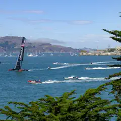 High-performance sailboats race across a bay, surrounded by smaller boats and a scenic shoreline under a clear sky.