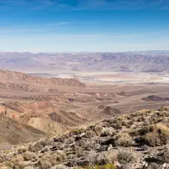 A rocky hillside with sparse vegetation leads to a vast desert valley surrounded by rugged mountains under a clear blue sky.