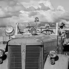 Two skeletons wearing firefighter helmets sit in the cab of a vintage fire truck, parked in a desert landscape with rocky formations in the background.