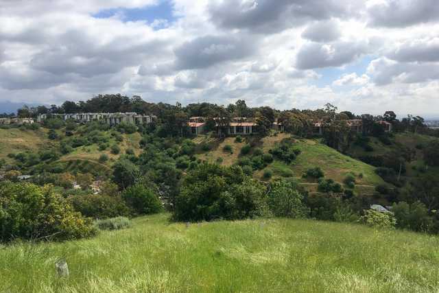 A hilly landscape with a cluster of houses on top and lush greenery below under a partly cloudy sky.