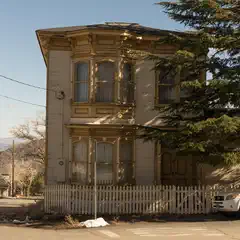 Two-story wooden house with yellow trim and white picket fence, near a street with a parked vehicle, against a mountainous backdrop.