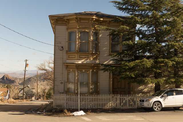 Two-story wooden house with yellow trim and white picket fence, near a street with a parked vehicle, against a mountainous backdrop.