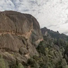 A rugged, layered rock formation rises above a forested valley under a partly cloudy sky.