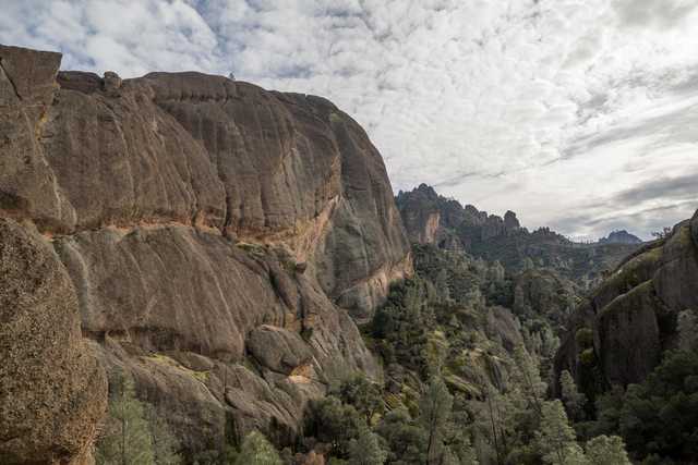 A rugged, layered rock formation rises above a forested valley under a partly cloudy sky.