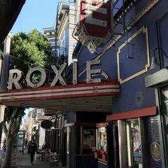 A vintage theater with a bold red and blue facade and a prominent marquee featuring the name "ROXIE" is situated on a city street.