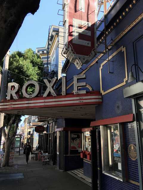 A vintage theater with a bold red and blue facade and a prominent marquee featuring the name "ROXIE" is situated on a city street.