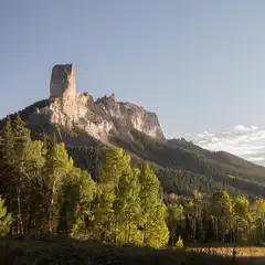 A towering rocky peak rises above a forested valley under a clear sky.