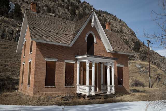 An old red brick building with white trim and a porch supported by columns stands alone in front of a mountain range.