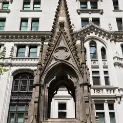 A Gothic-style stone monument with a pointed arch and decorative spire stands in front of a multi-story building with ornate windows and classical detailing.