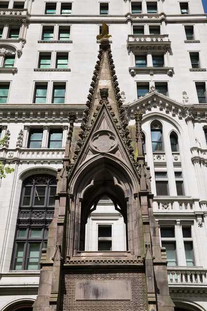 A Gothic-style stone monument with a pointed arch and decorative spire stands in front of a multi-story building with ornate windows and classical detailing.
