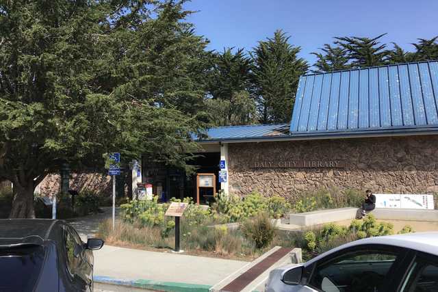 A building with a blue roof and stone walls, surrounded by trees and plants, with cars parked nearby on a paved area.