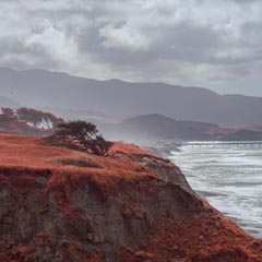 A red-tinged cliff overlooks a rocky coastline with a long pier extending into the ocean under an overcast sky.