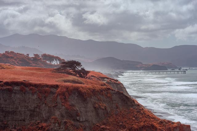 A red-tinged cliff overlooks a rocky coastline with a long pier extending into the ocean under an overcast sky.