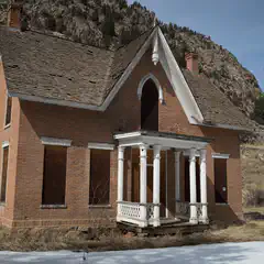 An old red brick building with white trim and a porch supported by columns stands alone in front of a mountain range.