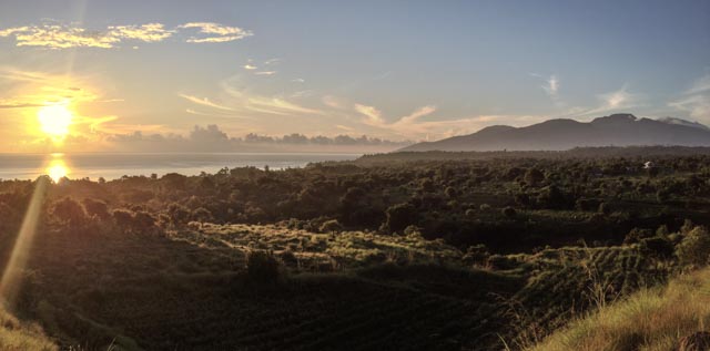 Sunset over calm water with distant hills, hazy clouds, and silhouetted trees; serene landscape features warm hues under fading light.