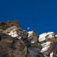 A rocky cliff face under a clear blue sky with the moon visible above it.