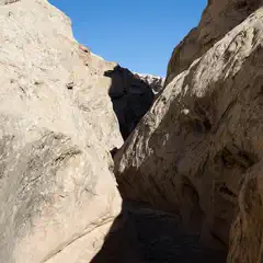 A narrow slot canyon with high, light-colored rock walls and a sandy floor.